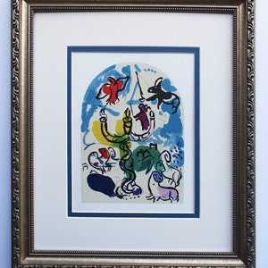 Op de afbeelding: Een ingelijste kunstdruk met een kleurrijk abstract ontwerp. Het kunstwerk bevat een gele kandelaar, rode vogels en figuren in blauwe, groene en paarse tinten. De print is geplaatst in een witte passe-partout en een goudkleurige, sierlijke lijst.