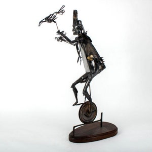 May include: A metal sculpture of a person riding a unicycle and holding a bird in their hand. The sculpture is made of recycled metal and has a rustic, industrial look.