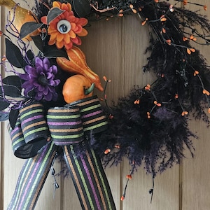 Può includere: Una ghirlanda di Halloween con base nera, adornata con elementi arancioni e viola. Presenta zucche decorative, un grande fiocco a righe e un fiore con un occhio. Perfetta per l'arredamento stagionale.