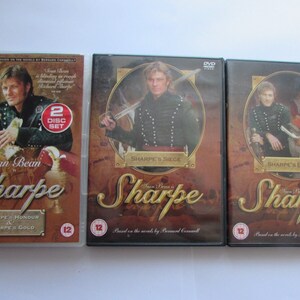 May include: Three DVD cases for the Sharpe series starring Sean Bean. The cases are red, black, and gold with the title "Sharpe" in a stylized font. The text "Sean Bean is Sharpe" is also on the cases. The cases are labelled with the episode titles "Sharpe's Siege", "Sharpe's Enemy", and "Sharpe's Honour & Sharpe's Gold".