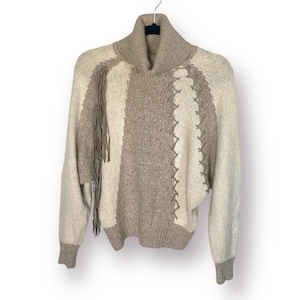 Puede incluir: Un suéter de punto beige y blanco con cuello alto y detalle de flecos. El suéter tiene un patrón geométrico y está hecho de un material suave y acogedor.