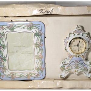 May include: A decorative porcelain photo frame and clock set. The frame is light blue with floral accents and a white center. The clock has a gold-tone face and Roman numerals. The set is in a box with the brand name "Kirch".
