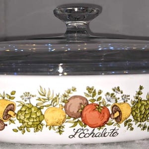 May include: A white oval ceramic casserole dish with a clear glass lid and a knob handle. The exterior features a colorful botanical illustration of vegetables including mushrooms, tomatoes, and artichokes, with the text L'Echalote printed below.