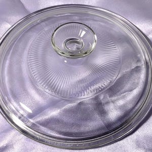 Könnte beinhalten: Ein klarer Pyrex-Glasdeckel mit einem zentralen Griff. Der Deckel hat ein kreisförmiges Design mit vertikalen Rippen, die von der Mitte ausgehen. Der Griff ist ein kleiner, abgerundeter Knopf. Der Deckel ist für eine runde Schüssel konzipiert.