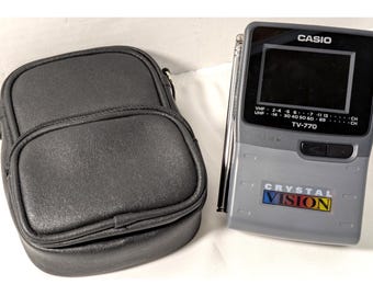 May include: A vintage Casio TV-770 portable television with a small screen, antenna, and a black leatherette carrying case. The TV is gray with the words "CASIO" and "CRYSTAL VISION" printed on it.