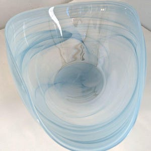 May include: A light blue and white glass bowl with a unique, asymmetrical shape. The bowl features a swirling pattern of light blue and white, creating a marbled effect. The glass is translucent, allowing light to pass through, and the bowl has a smooth, polished finish.