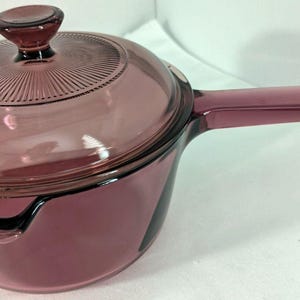 May include: A vintage, amethyst-colored saucepan with a matching lid. The glass lid has a ridged design and a small knob. The saucepan features a long, integrated handle and a pouring spout. This cookware is ideal for cooking and serving.