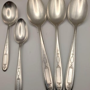 May include: A set of five silver-toned metal spoons of varying sizes arranged vertically. Each spoon features a slender handle with a decorative beaded border, an elongated rectangular panel near the base, and intricate floral-inspired detailing.