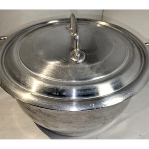 May include: A silver aluminium pot with a lid and two handles. The pot is round and has a smooth surface.