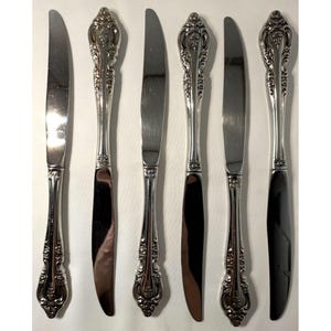 Könnte beinhalten: Sechs silberfarbene Messer mit verzierten, detaillierten Griffen sind in einer Reihe angeordnet. Die Klingen sind glatt und reflektierend. Die Griffe weisen ein florales Design auf. Die Messer sind wahrscheinlich für formelle Anlässe gedacht.