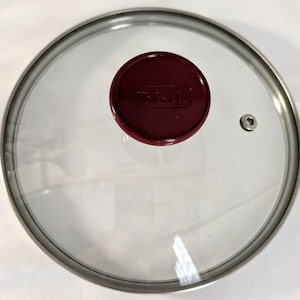 Puede incluir: Una tapa circular de vidrio transparente para olla o sartén, con un borde de metal plateado y un pomo redondo de color granate oscuro en el centro. El nombre de la marca T-fal está grabado en el pomo y se observa una pequeña válvula de vapor.