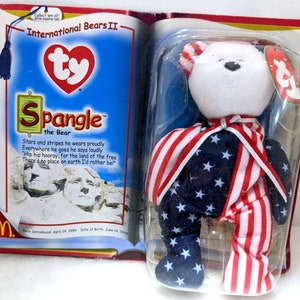 May include: A white Ty Beanie Baby bear wearing a red, white, and blue outfit with stars and stripes. The bear is packaged in a plastic case with a red, white, and blue background. The text "Spangle the Bear" is printed on the packaging. The text "International Bears II" is also printed on the packaging.