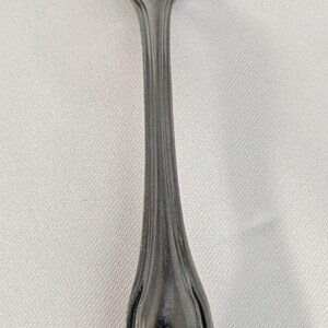 Peut inclure: Une cuillère argentée brillante avec un bol arrondi et un long manche fin. La poignée présente un design décoratif à nervures et des motifs floraux à la base. Cet ustensile est probablement destiné à servir ou à manger.