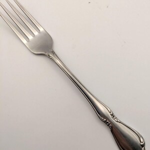 May include: A silver-toned metal dinner fork featuring four tines and a slender handle. The handle is decorated with embossed floral motifs at the neck and a rounded, flared base with additional small flower detailing at the very bottom edge.