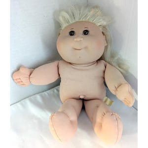 May include: A vintage Cabbage Patch Kid doll with a soft, fabric body and limbs. The doll has a round face with rosy cheeks, dark eyes, and a small smile. Its light blonde hair is styled in a soft, wavy fashion.