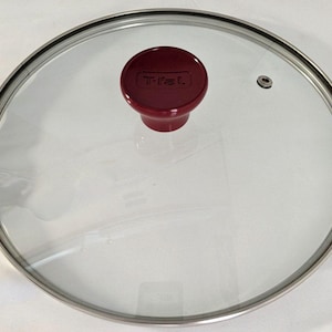 May include: A circular tempered glass lid with a stainless steel rim and a dark red knob handle. The knob features the embossed T-fal logo. A small circular steam vent is integrated into the glass surface, positioned near the outer edge.