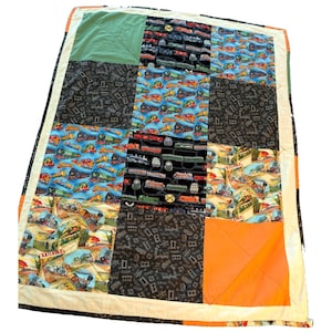 Puede incluir: Un edredón de retazos con temática de trenes. El edredón incorpora cuadrados de tela azul, negra y naranja, con patrones de trenes y ferrocarriles. El edredón está bordeado con una tela de color crema y tiene un cuadrado verde en la esquina superior izquierda.