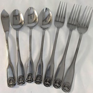 May include: A set of stainless steel silverware including a butter knife, spoons, and forks. The silverware has a decorative shell design on the handle. The utensils are arranged on a white surface.