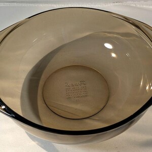 May include: A round, translucent brown glass mixing bowl with integrated side handles. The interior base features embossed text that reads Pyrex, Made in USA, and other manufacturing markings. The bowl has a smooth, curved design.
