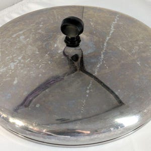 Può includere: Un coperchio circolare in metallo con una superficie argentata lucida e riflettente che mostra segni di ossidazione e usura. Il coperchio presenta un pomello centrale nero di forma cilindrica con una parte superiore arrotondata.