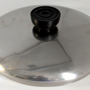 May include: A circular, polished stainless steel pot lid with a reflective surface. The lid features a centered, black, circular knob with a slightly recessed top, providing a secure grip for lifting the lid from a cooking vessel.