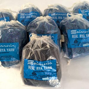 Könnte beinhalten: Mehrere Beutel mit blauem und schwarzem Garn werden gezeigt. Jeder Beutel hat ein blaues Etikett mit dem Text "Spinnerin REAL RYA YARN". Das Garn ist in transparenten Plastiktüten verpackt und mit weißer Schnur gebunden.