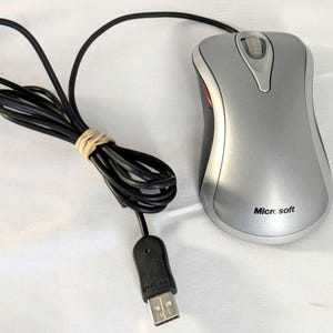 May include: A silver and black wired computer mouse with a scroll wheel and the Microsoft logo printed on the top surface. The device includes a black USB cable that is coiled and secured with a rubber band, ending in a standard USB connector.
