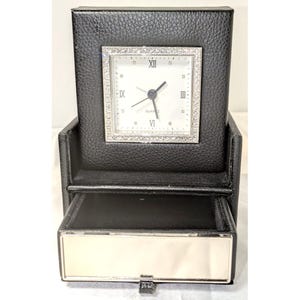 May include: A black leather desk clock with a silver face and Roman numeral hour markers. The clock has a small drawer underneath.
