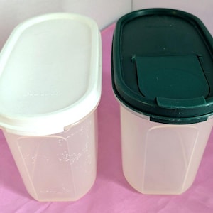 May include: Two clear plastic food storage containers with oval-shaped lids. One container has a white lid, and the other has a dark green lid. The containers are translucent, allowing the contents to be seen. The lids have a secure closure.