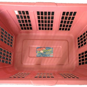 May include: A rectangular, light pink laundry basket with multiple rectangular ventilation openings. The basket has a smooth interior and a slightly textured exterior. A small, colorful sticker is visible on the inside bottom.