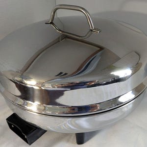 May include: A stainless steel electric buffet server with a domed lid and side handles. The server sits on a black base with short legs. The lid has a curved handle for easy lifting. The server is designed to keep food warm.