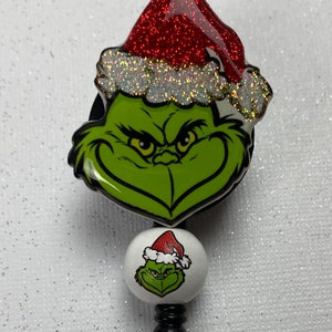 May include: A green Grinch face with a red glitter Santa hat and a white glitter trim. The Grinch is on a black retractable badge reel with a smaller Grinch image on the white button.