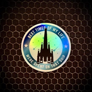 May include: A round sticker with the text "BEST TIMES OF MY LIFE WERE SPENT ON THAT SHIP" encircling a black spaceship silhouette. The sticker has a holographic, iridescent finish with blue and green hues, and is set against a dark, textured background.