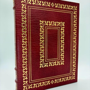 May include: A hardcover book with a deep red cover and gold decorative accents. The cover features a gold border and floral-like corner designs. The spine is also red with gold lettering. The book is set against a white background.