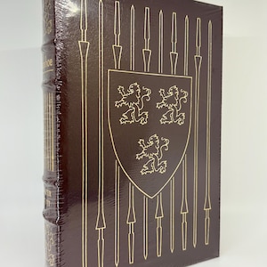May include: A hardcover book wrapped in clear plastic. The cover is dark brown with gold-colored illustrations of spears and a shield with three lion emblems. The spine has gold lettering.
