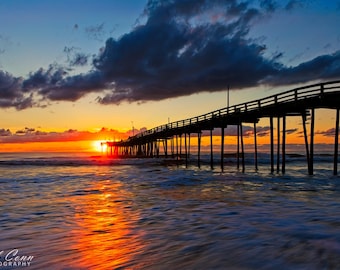 May include: A scenic photograph of a pier extending into the ocean at sunset. The sky transitions from blue to orange and yellow, with dark clouds overhead. The water reflects the warm colors of the setting sun.
