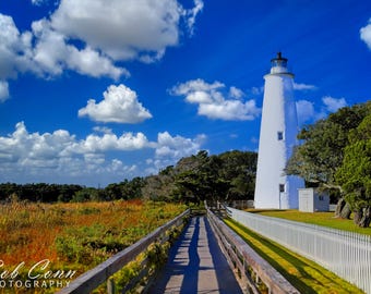 May include: A tall, white lighthouse stands against a bright blue sky dotted with fluffy white clouds. A wooden walkway leads towards the lighthouse, with a white picket fence on the right. The foreground features a field of tall grass.