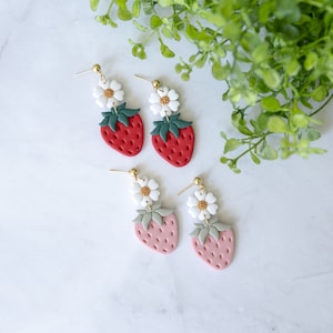 以下が含まれることがあります： イチゴと白い花のモチーフがついた、ぶら下がりのイヤリングです。ポリマークレイで作られており、金色の仕上げが施されています。