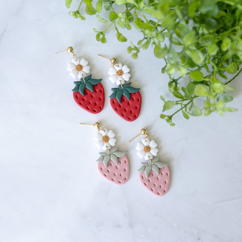 以下が含まれることがあります： イチゴと白い花のモチーフがついた、ぶら下がりのイヤリングです。ポリマークレイで作られており、金色の仕上げが施されています。