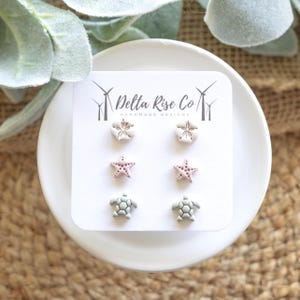 May include: A white card displays six pairs of handmade earrings. The earrings are in the shape of a flower, starfish, and turtle. The flower earrings are white, the starfish are pink, and the turtles are green. The card has the text "Delta Rise Co HANDMADE DESIGNS".