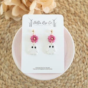 May include: A pair of dangle earrings featuring a white ghost charm with black eyes and mouth suspended below a dark pink five-petaled flower with a gold center. The earrings are attached to a white display card labeled Delta Rise Co.