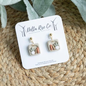 May include: A pair of beige clay earrings with gold hooks. Each earring features a miniature bookshelf with a small potted plant and books. The earrings are displayed on a white card with the text "Delta Rise Co Handmade Designs".