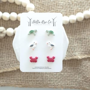 May include: A white card displays six pairs of stud earrings with a coastal theme. The earrings feature turtles, seagulls, and crabs in green, white, gray, and pink. The card has the text "Delta Rise Co" and "Handmade Designs".