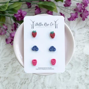 May include: A set of six handmade stud earrings in the shape of strawberries, blueberries, and raspberries. The earrings are red, blue, and pink, with green accents. The earrings are displayed on a white card with the text "Delta Rise Co Handmade Designs".