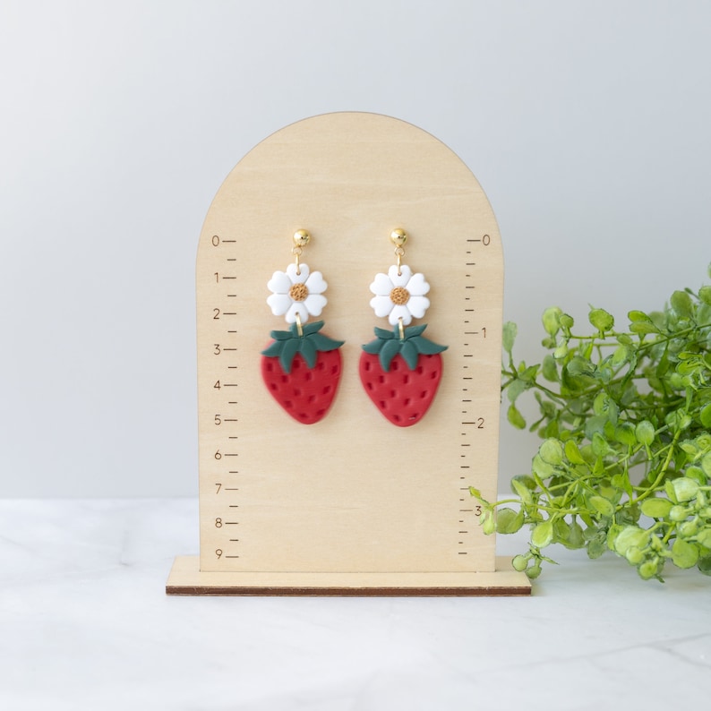 以下が含まれることがあります： 赤いイチゴと緑の葉、白い花がついた、ぶら下がりのイヤリングのペア。イヤリングは、定規が付いた木製のスタンドに飾られています。