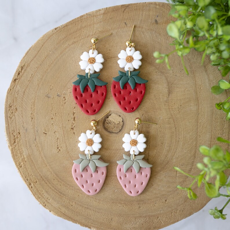 以下が含まれることがあります： 白い花のチャームと赤いイチゴのチャームが付いた、ぶら下がりのイヤリングのペア。イヤリングはポリマークレイ製で、金色の金具が付いています。