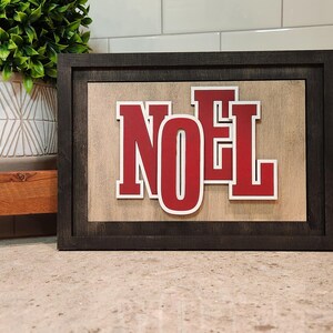 May include: A framed wooden sign with the word "NOEL" in red letters with white outlines. The sign has a dark gray frame and a light brown background. The sign is a decorative item.