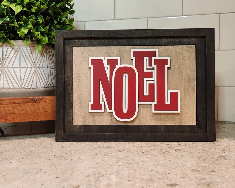 May include: A framed wooden sign with the word "NOEL" in red letters with white outlines. The sign has a dark gray frame and a light brown background. The sign is a decorative item.