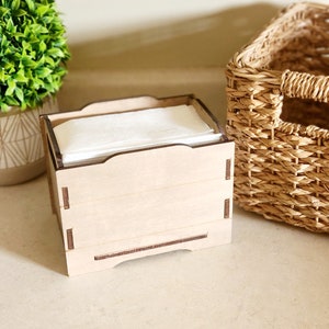May include: A wooden tissue box holder with a natural wood finish. The holder is empty and has a rectangular shape with rounded edges. The holder is sitting on a light brown surface.
