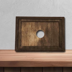 May include: A rectangular wooden board with a dark brown frame and a lighter brown center. A circular hole is cut in the center of the board. The board is displayed on a wooden surface.